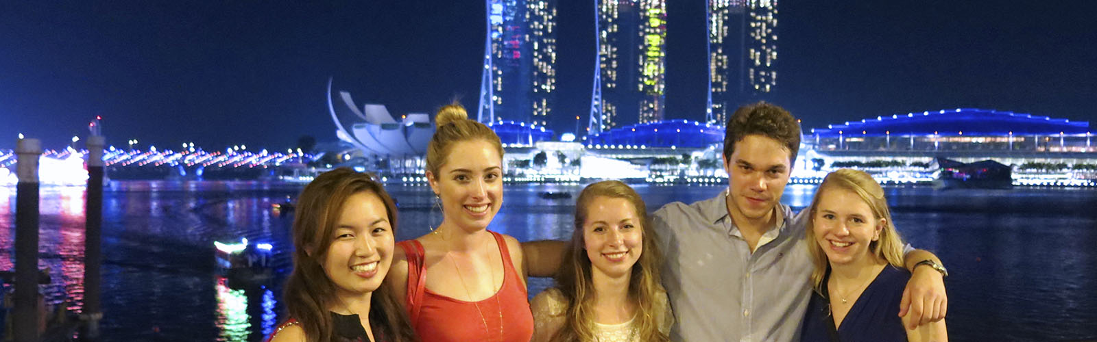 Five students posed on a waterfront walkway at night, with the brightly-lit Marina Bay Sands towers in the background. 